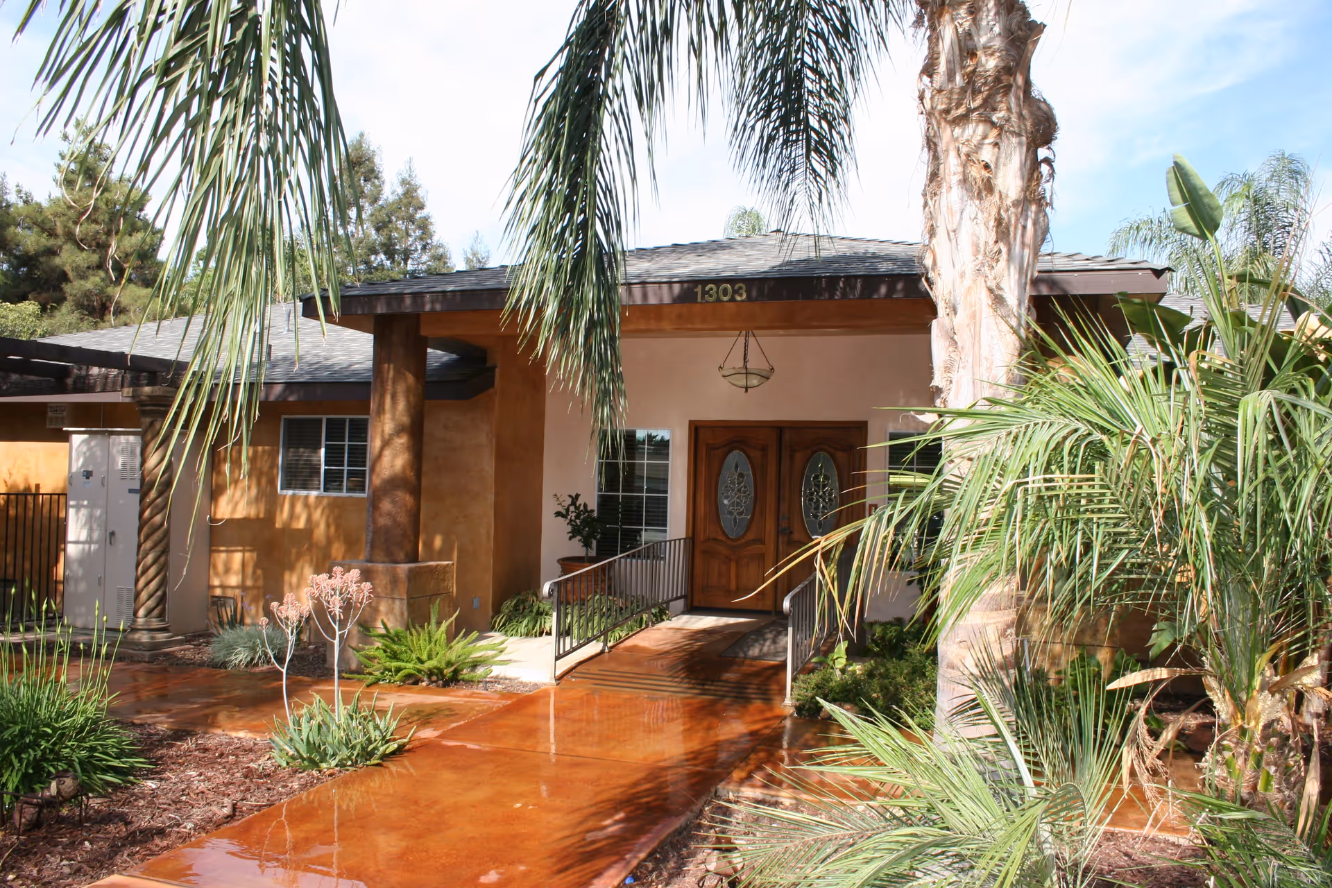Front entrance of a building with double wooden doors featuring decorative glass panels, a covered porch supported by two columns, a hanging light fixture, and a wet reddish-brown concrete walkway surrounded by various green plants and palm trees.
