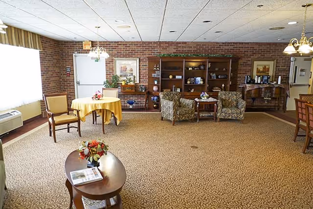 A spacious common area with patterned carpet, two upholstered armchairs around a small table, a wooden bookshelf with decorative items, a round table covered with a yellow tablecloth and two chairs, a wooden coffee table with a flower arrangement and a magazine, and brick walls with framed artwork and a door.