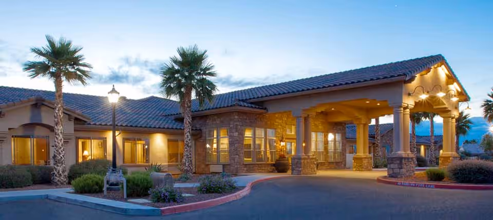 Exterior view of Rock Creek at The Park Memory Care Community building at dusk, featuring a covered entrance with stone pillars, warm lighting, palm trees, and landscaped bushes along the driveway.