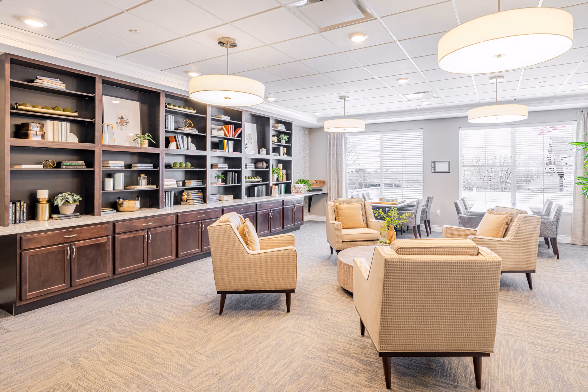 A bright and spacious living room area with four beige armchairs arranged around a round wooden coffee table. Behind the seating area is a large built-in dark wood bookshelf filled with books, plants, and decorative items. Large windows with blinds allow natural light to fill the room, and there are two dining tables with chairs near the windows. The ceiling has recessed lighting and three modern round pendant lights.