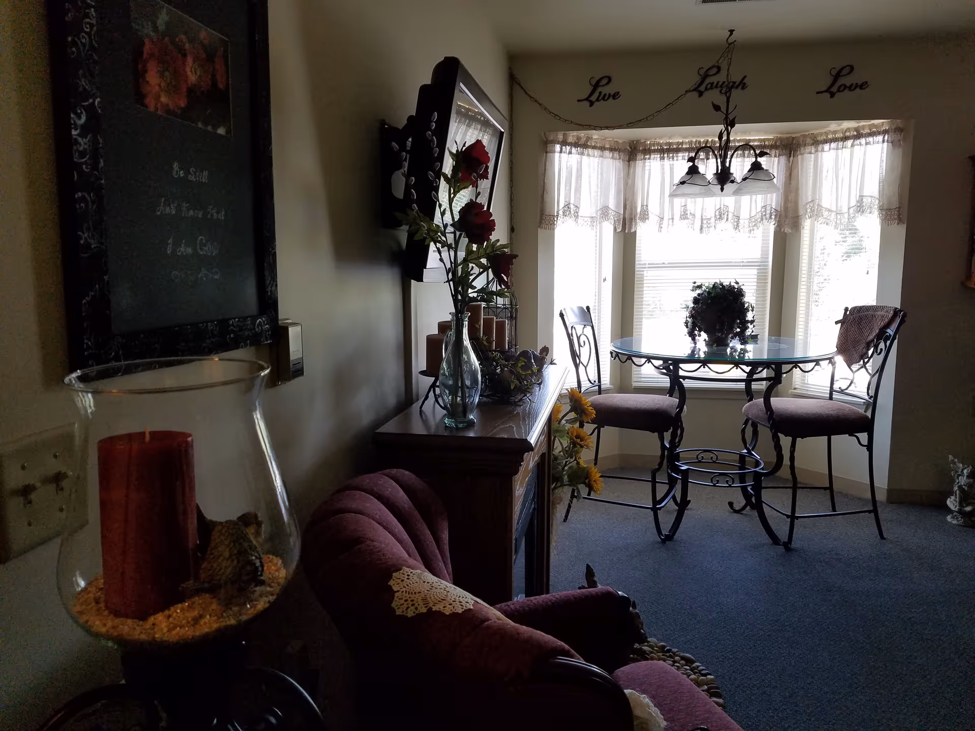 A cozy interior corner of a room with a small round glass table and two chairs near a window with lace curtains. Above the window, the words 'Live Laugh Love' are displayed on the wall. There is a burgundy upholstered chair with a doily on the armrest, a wooden cabinet with decorative items including a vase with red flowers, and a candle in a glass holder in the foreground.