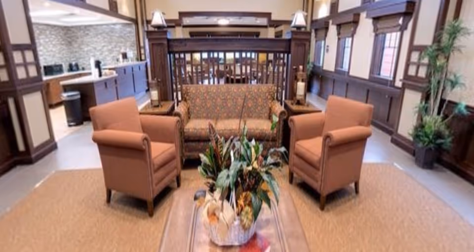 A furnished common seating area with a patterned sofa, two armchairs, and a coffee table topped with plants in a well-lit interior space.