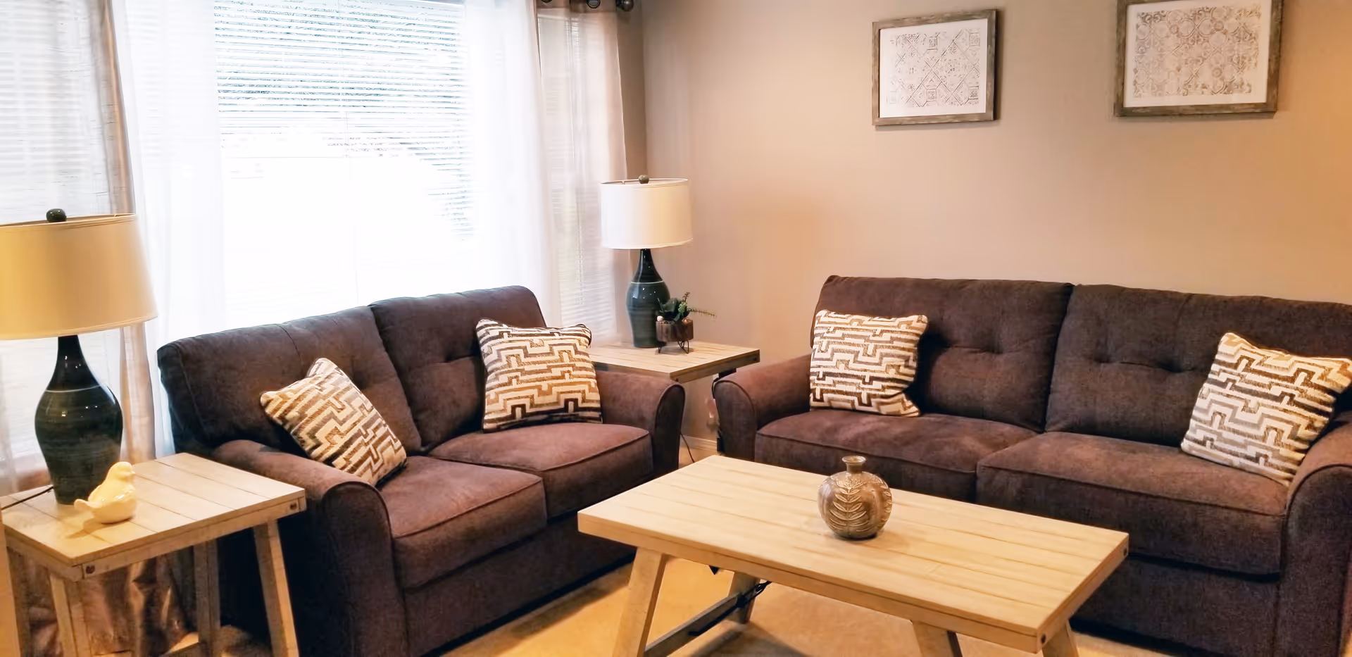 A cozy living room with two dark brown sofas, each adorned with patterned throw pillows. There are two wooden side tables with lamps on either side of the sofas, and a wooden coffee table in the center with a decorative vase. The room has light-colored walls with two framed artworks and large windows with sheer curtains allowing natural light to fill the space.