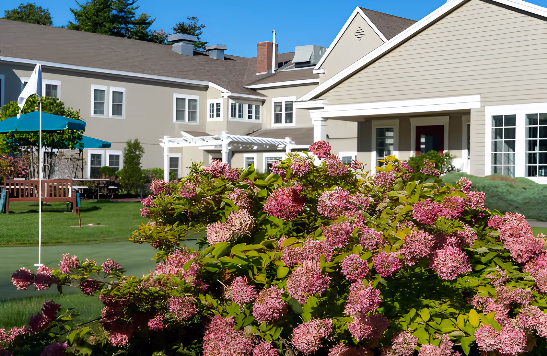 Exterior view of Granite Hill Estates showing a beige building with white trim, surrounded by green grass and vibrant pink flowering bushes under a clear blue sky. There is a white flag on a pole and a wooden bench near the building.