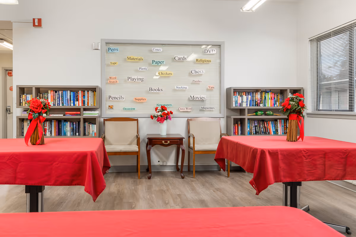 A well-lit room with tables covered in red tablecloths, each adorned with a vase of red flowers. Two beige chairs flank a small wooden table with a flower vase in the center. Behind the chairs is a whiteboard with various words related to activities and materials. Bookshelves filled with books are on either side of the chairs. A window with blinds is on the right wall.