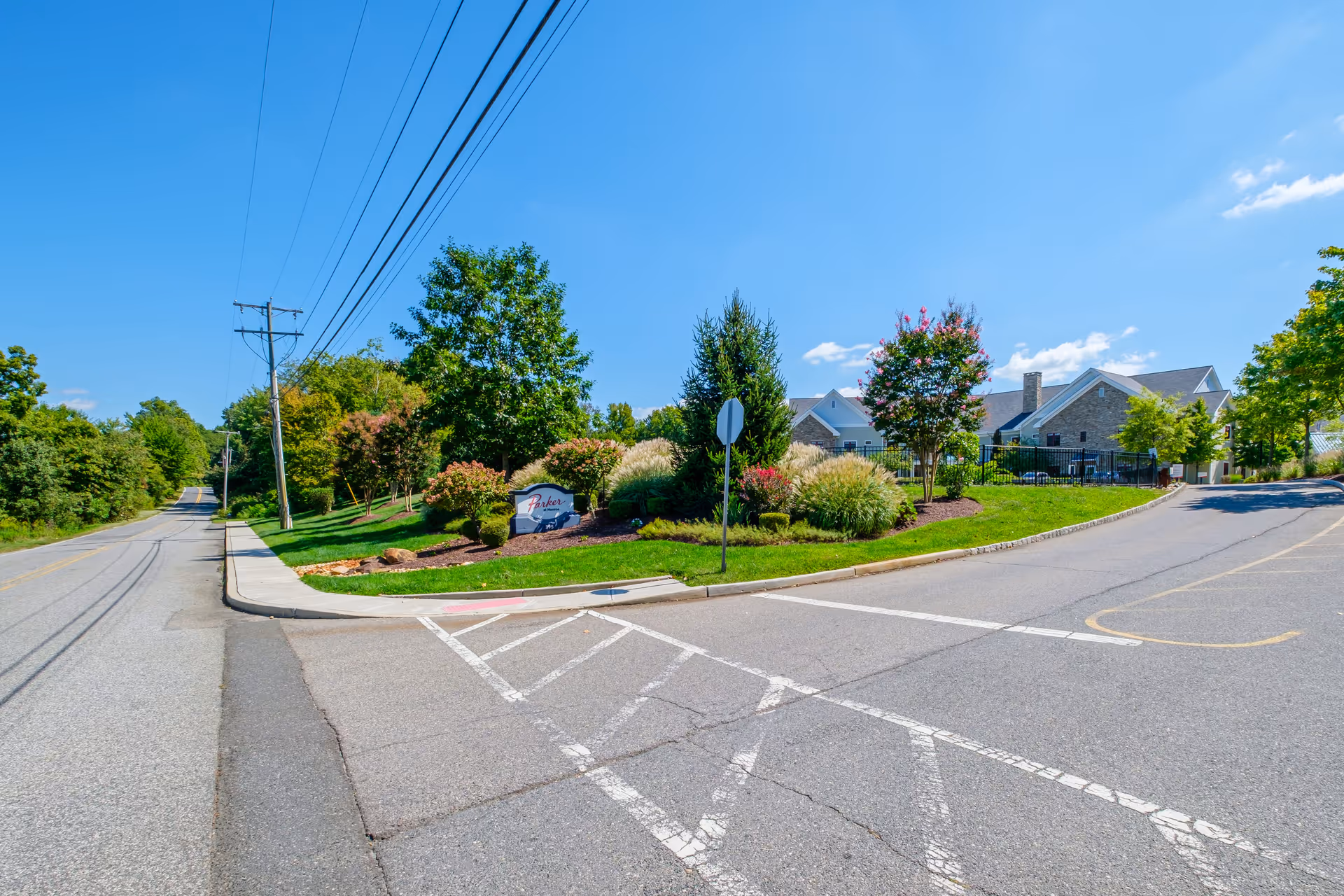Entrance to Parker at Monroe senior living facility with a landscaped area featuring trees, bushes, and a sign. The building is visible in the background under a clear blue sky.
