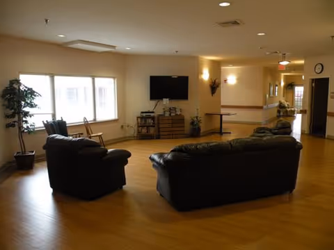 A spacious living area with wooden flooring, two black leather sofas facing a wall-mounted flat screen TV, a small wooden cabinet with books and decor, a potted plant near a large window with blinds, and a hallway leading to other rooms.