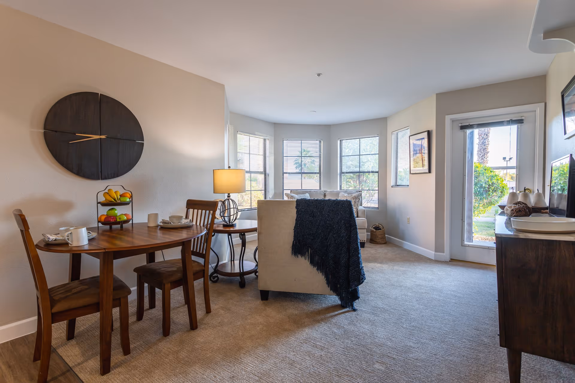 A cozy living room and dining area in a senior living facility. The dining area features a wooden table with two chairs, set with cups and plates, and a tiered fruit basket. A large round wall clock hangs above the table. The living room has a beige armchair with a dark throw blanket, a side table with a lamp, and a sofa near a bay window letting in natural light. A door with a window leads outside, and framed pictures hang on the walls.