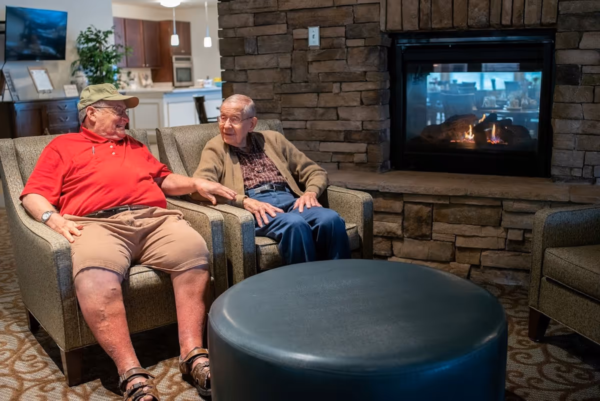 Two elderly men sitting and talking in a lounge area with armchairs and a stone fireplace.