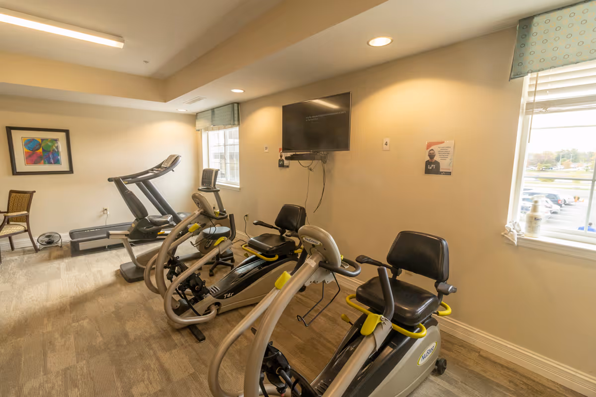 Small fitness room with recumbent exercise bikes, a treadmill, chairs, a wall-mounted TV, and windows.