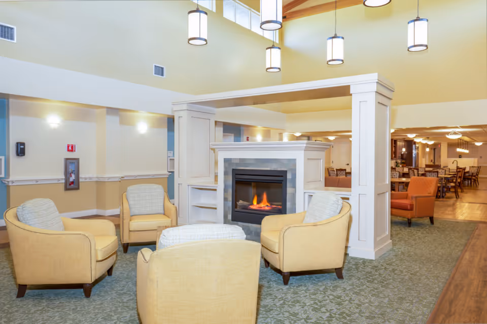 A bright communal lounge with four yellow armchairs arranged around a central fireplace and pendant lights, with a dining area visible in the background.