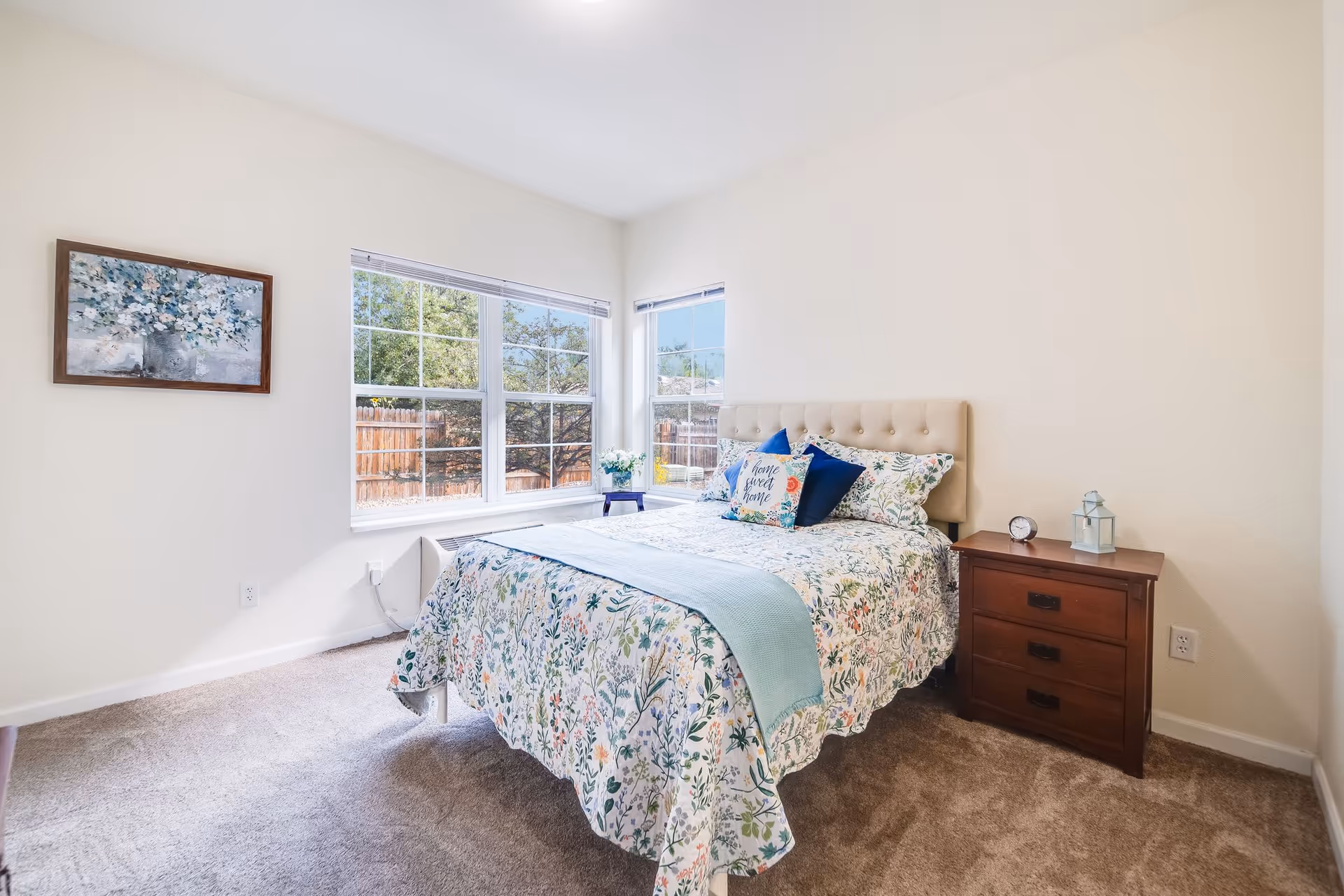 A bright bedroom with a bed covered in floral bedding and decorative pillows, including one that says 'home sweet home'. The room has beige walls, a carpeted floor, a wooden nightstand with a small clock and lantern, and large windows letting in natural light with a view of a fenced backyard.
