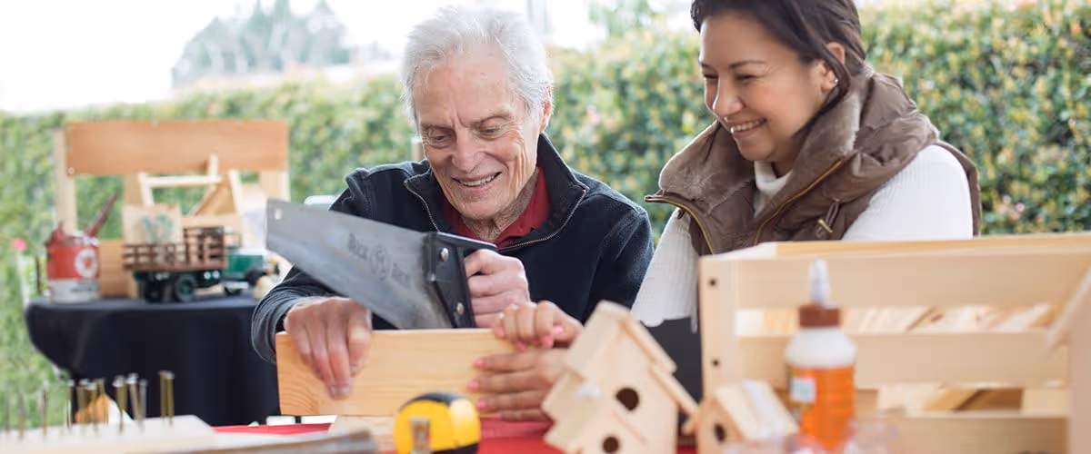 An elderly man and a younger woman are smiling and working together on a woodworking project outdoors. The man is using a hand saw to cut a piece of wood while the woman watches and supports the wood. Various woodworking tools and wooden items are visible on the table in front of them.