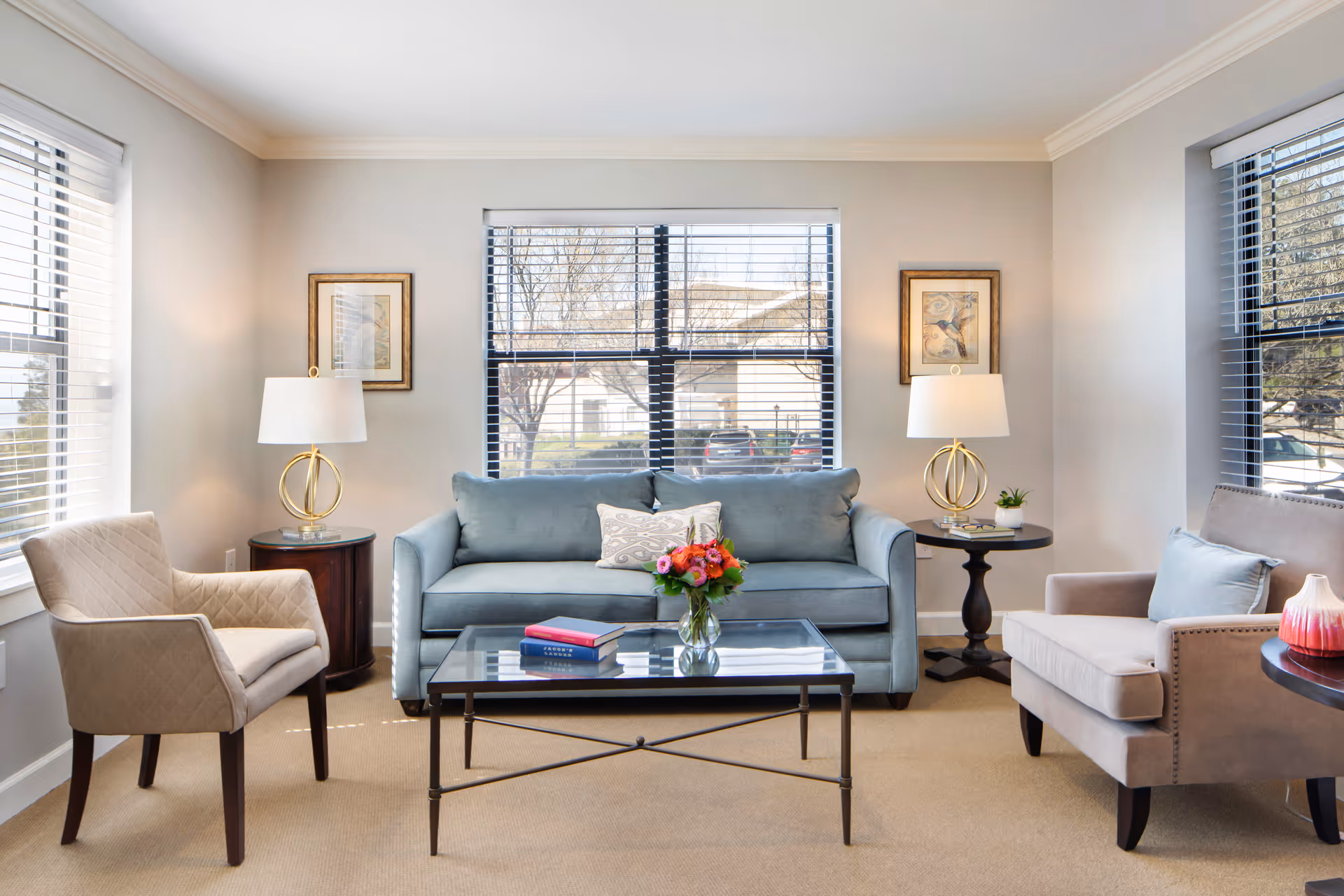 A bright and cozy living room with a light blue sofa centered between two windows with blinds. There are two beige armchairs on either side of the sofa, each next to a round side table with a lamp. A glass coffee table with a vase of flowers and two books is in front of the sofa. The walls are light-colored with two framed pictures hanging above the side tables.