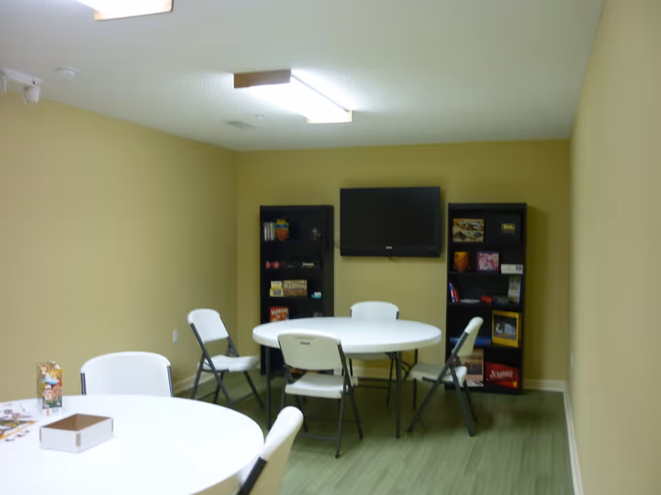 A small room with beige walls and green flooring featuring two round white tables surrounded by white folding chairs. Against the far wall, there are two black bookshelves filled with books and games, and a flat-screen TV mounted on the wall above them. The room is lit by a rectangular ceiling light.