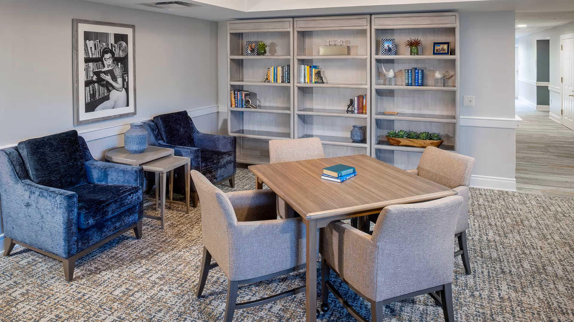 A cozy common area with a square wooden table surrounded by four beige upholstered chairs. Behind the table is a large bookshelf filled with books and decorative items. To the left, there are two blue velvet armchairs with a small side table between them. A black and white framed photograph hangs on the wall above the armchairs. The room has carpeted flooring and a hallway is visible in the background.