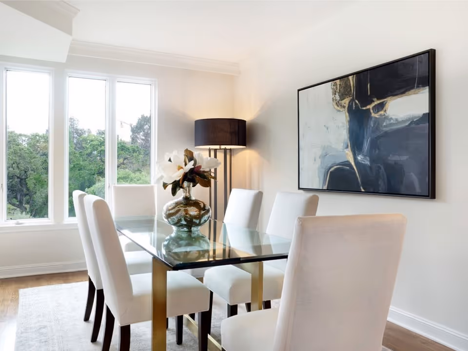 Bright modern dining room with a glass table surrounded by six upholstered chairs, a decorative vase centerpiece, floor lamp, and large abstract painting.