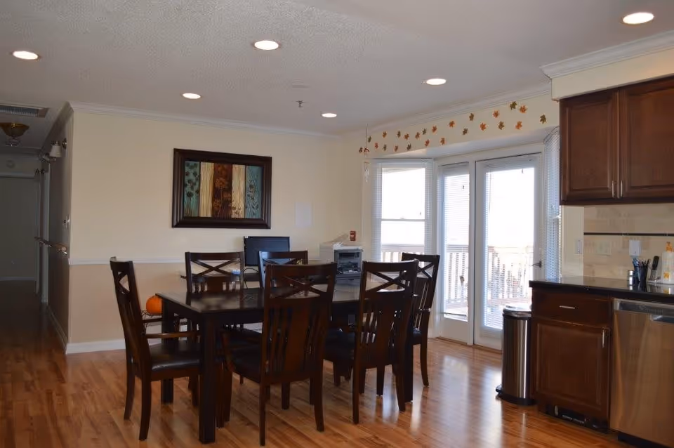 A dining area with a dark wooden table and six matching chairs on a polished wooden floor. The room has beige walls with a framed artwork hanging on one wall. There are double glass doors with blinds leading outside, and part of a kitchen with dark wooden cabinets and stainless steel appliances is visible on the right. The ceiling has recessed lighting.