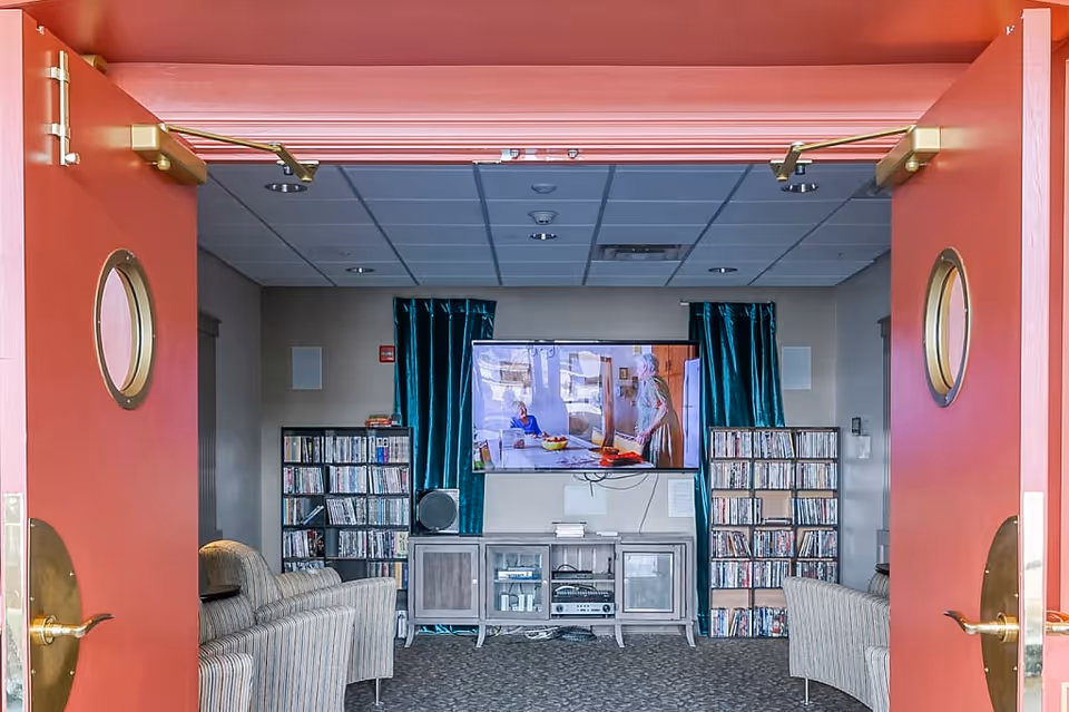 View through open double red doors into a cozy media room with a large flat-screen TV mounted on the wall, flanked by two tall shelves filled with DVDs. There are two striped armchairs facing the TV, a media console below the TV, and teal curtains on either side of the TV. The room has a carpeted floor and a drop ceiling with recessed lighting.