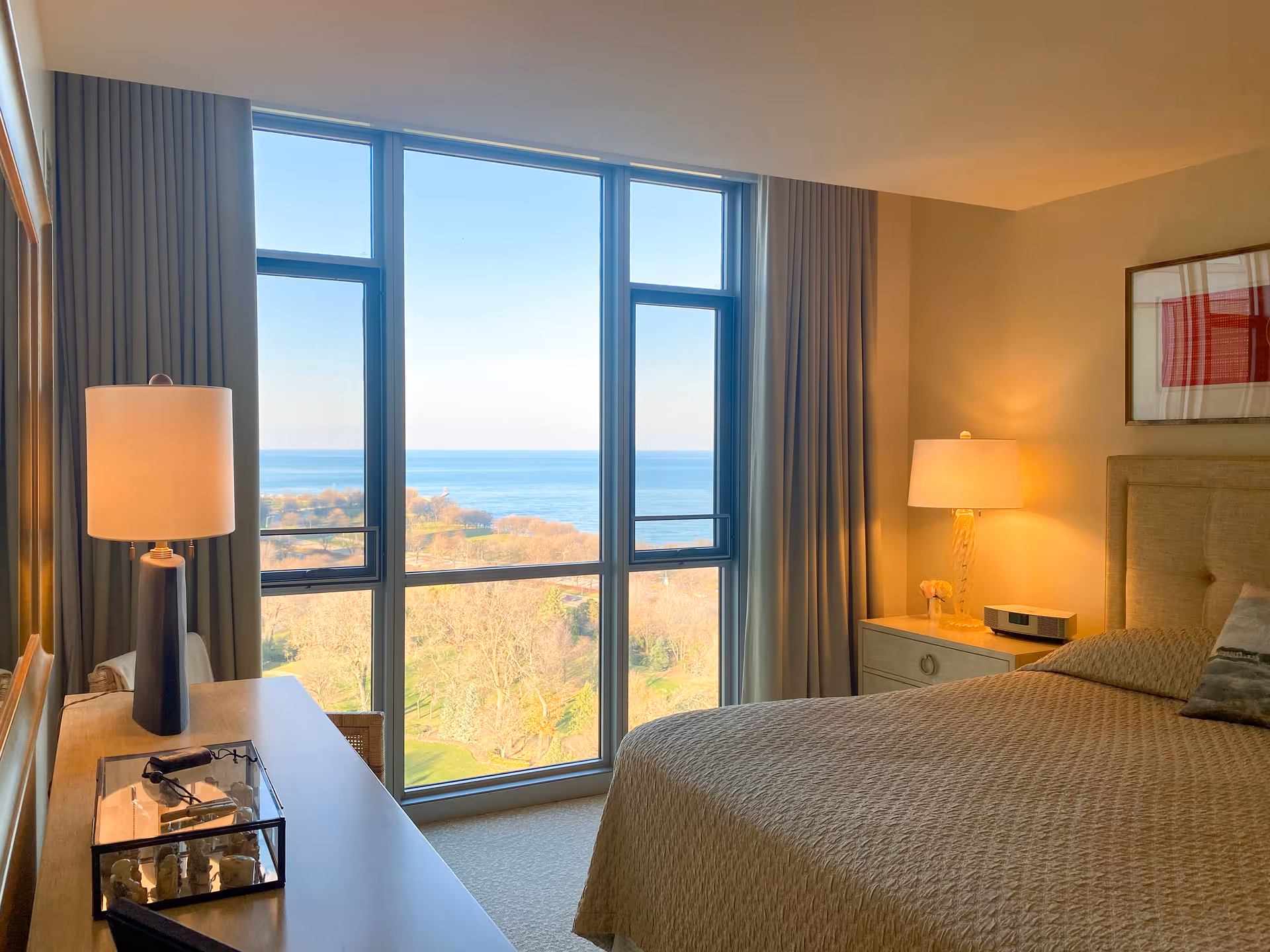 A cozy bedroom with a large window offering a scenic view of trees and a body of water. The room features a bed with a beige quilted bedspread, a nightstand with a lamp and a small speaker, a wooden dresser with a lamp and decorative items, and curtains framing the window.