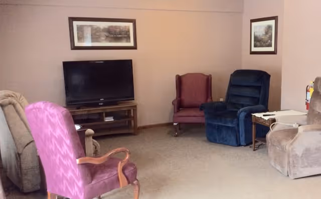 A cozy living room with a flat-screen TV on a wooden stand, surrounded by four armchairs in different colors including purple, blue, beige, and brown. The walls are light beige with two framed pictures hanging. There is a small side table with a white cloth and a fire extinguisher mounted on the wall.