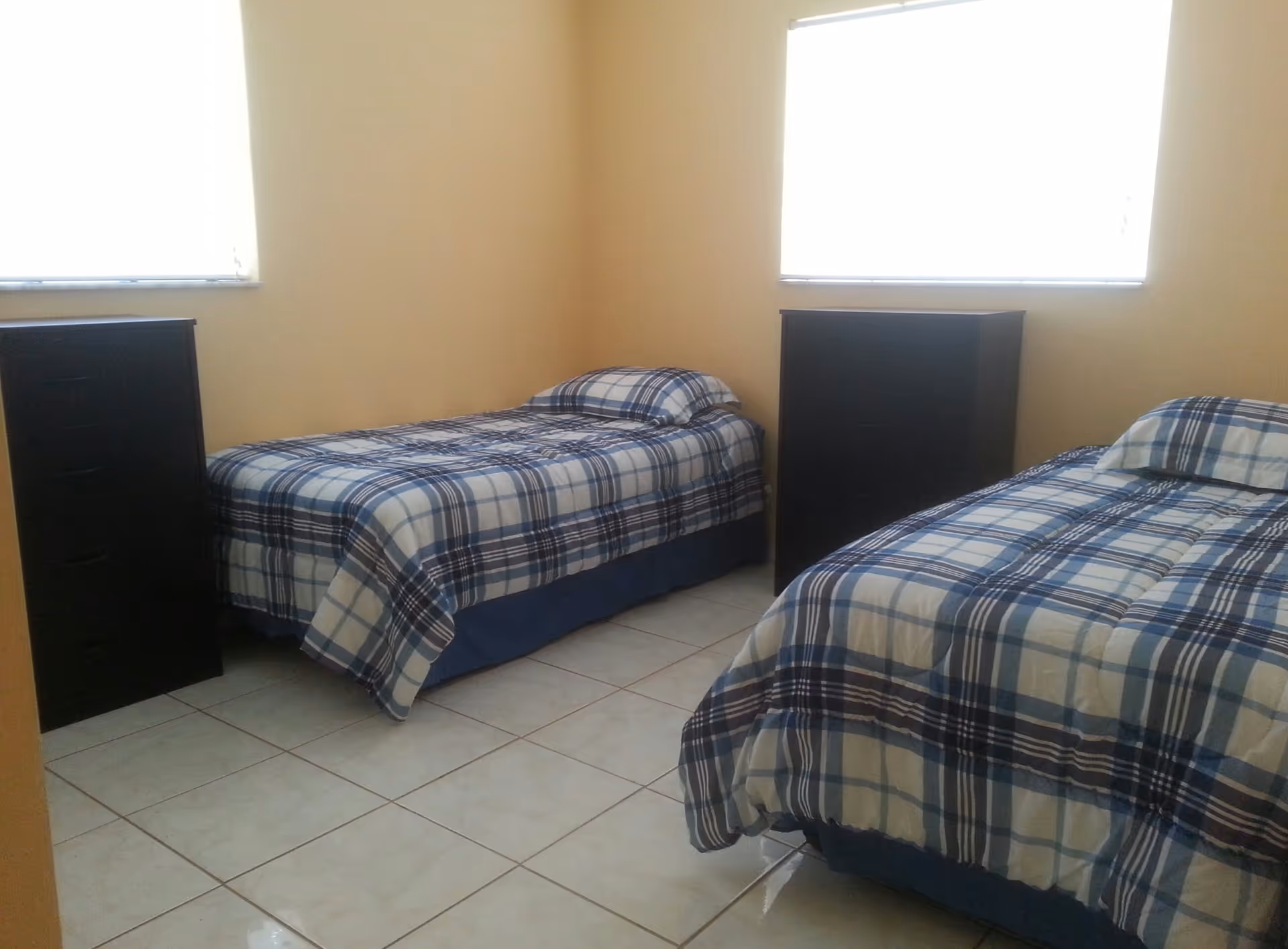 Small bedroom with two twin beds in blue plaid bedding, two dark dressers, tiled floor and two bright windows.