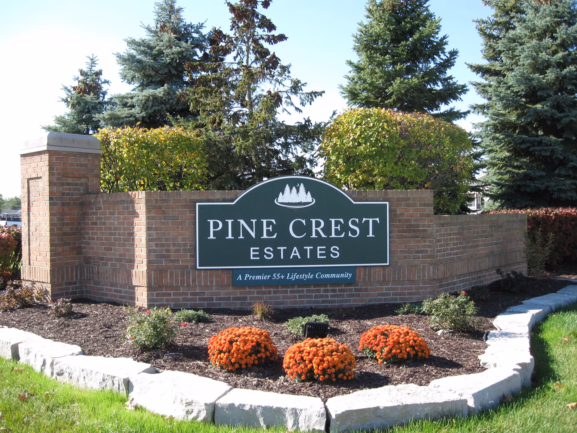 Brick entrance sign for Pine Crest Estates, a premier 55+ lifestyle community, surrounded by landscaping with orange flowers, green bushes, and pine trees in the background under a clear sky.