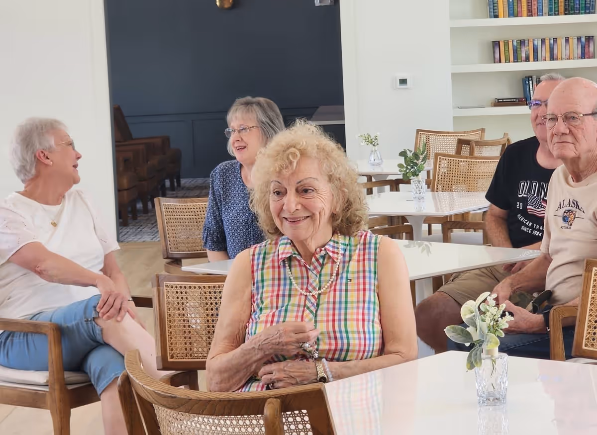 A group of elderly people sitting and chatting in a bright, modern common area with white tables, wooden chairs, and bookshelves in the background. Small vases with flowers are placed on the tables.
