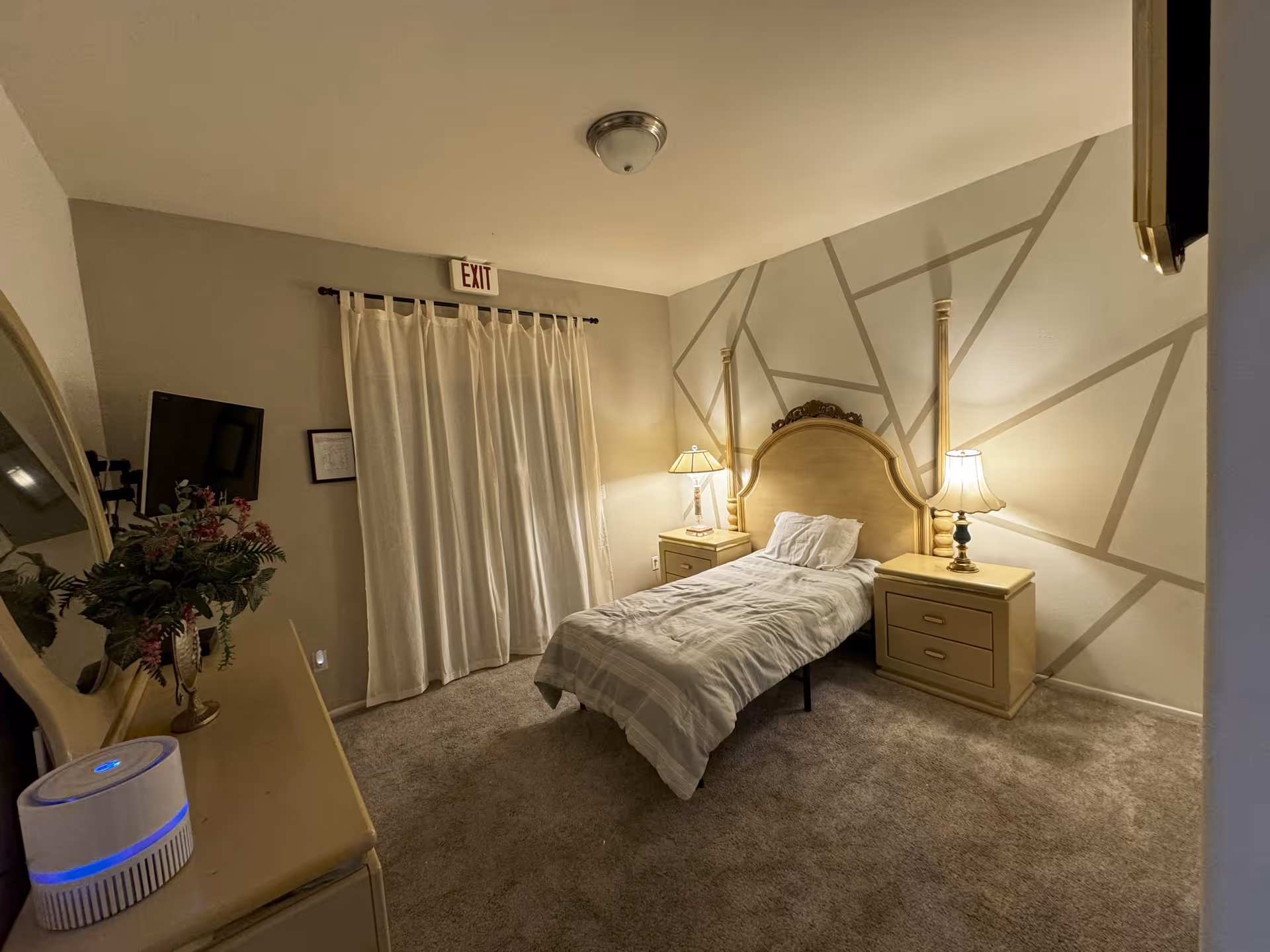 A cozy bedroom with a single bed centered against a wall with geometric patterns. The bed has a light-colored headboard and is flanked by two matching nightstands, each with a lamp. There is a dresser with a round mirror and a small plant on it. A curtain covers a window or door, and an exit sign is visible above the curtain. The room is softly lit and carpeted.