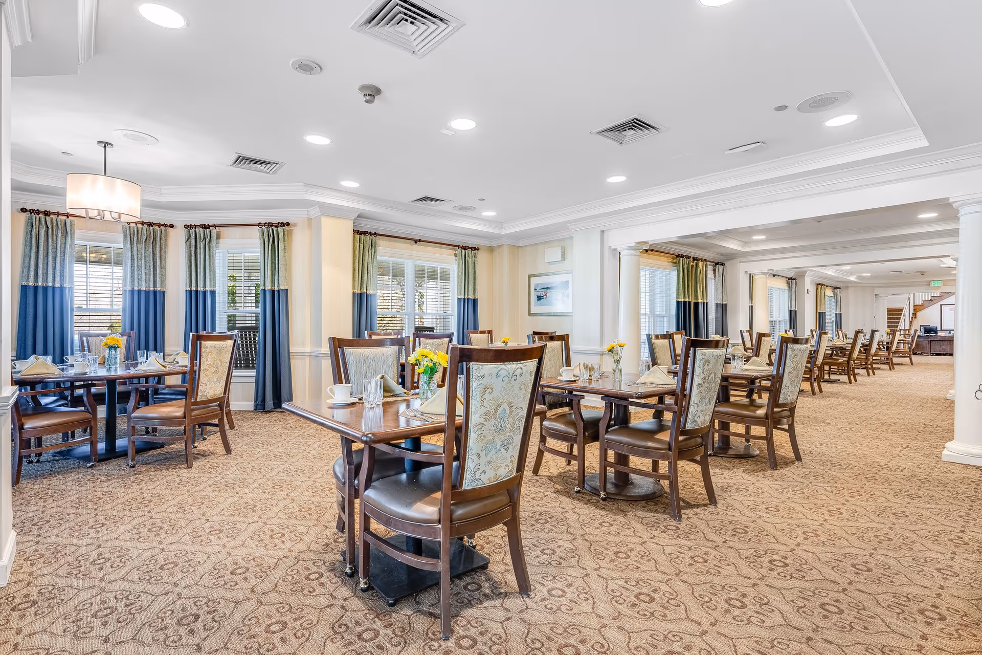 Bright, spacious dining room with multiple set tables and upholstered chairs, patterned carpet, and large windows with blue-and-green curtains.