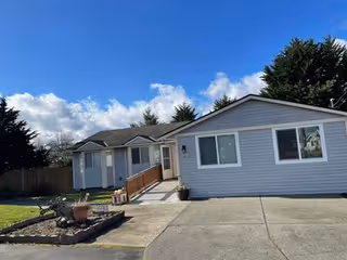 Single-story light gray building with white trim under a blue sky with some clouds. There is a concrete driveway and a small garden area with plants in front of the building. Trees are visible in the background.