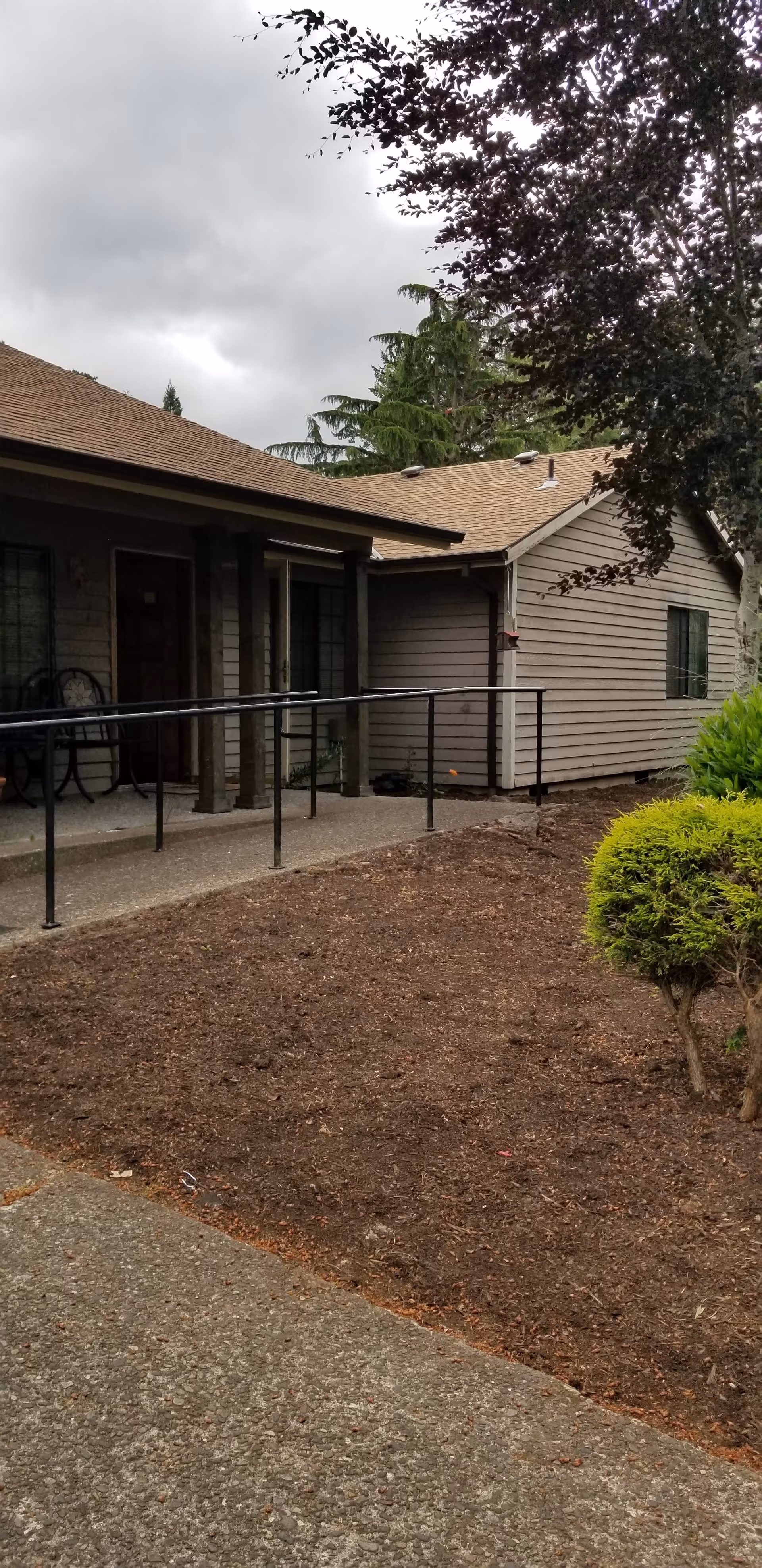 Exterior view of a single-story building with a brown roof and beige siding. There is a concrete walkway with a black metal handrail leading to a covered porch area with a door and windows. Trees and shrubs are visible around the building under a cloudy sky.