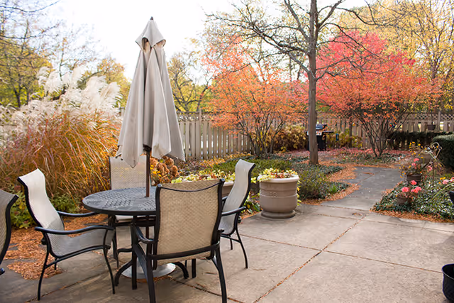 Outdoor patio area with a round metal table and four cushioned chairs surrounding it. A closed beige umbrella is positioned in the center of the table. The patio is surrounded by plants, trees with autumn-colored leaves, and a wooden fence in the background.