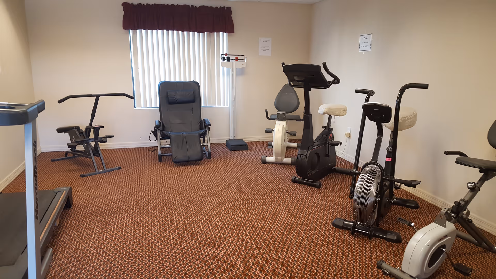 A small exercise room with various fitness equipment including a treadmill, rowing machine, stationary bikes, and a black recliner chair. The room has beige walls, a window with vertical blinds and a maroon valance, and a patterned carpeted floor.
