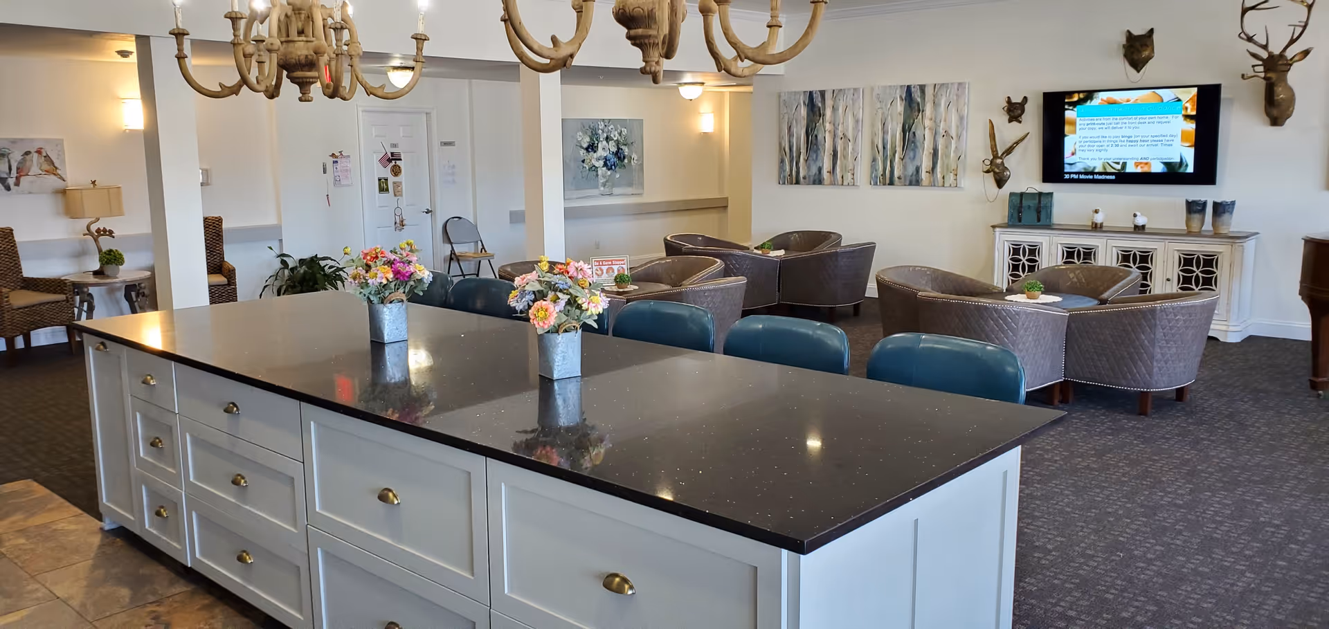 A senior living facility common area with a large kitchen island featuring a dark countertop and white cabinetry. The island has three small flower arrangements on top and several blue chairs along one side. In the background, there are multiple seating areas with wicker chairs and small tables, a wall-mounted TV, decorative wall art, and a chandelier hanging from the ceiling.