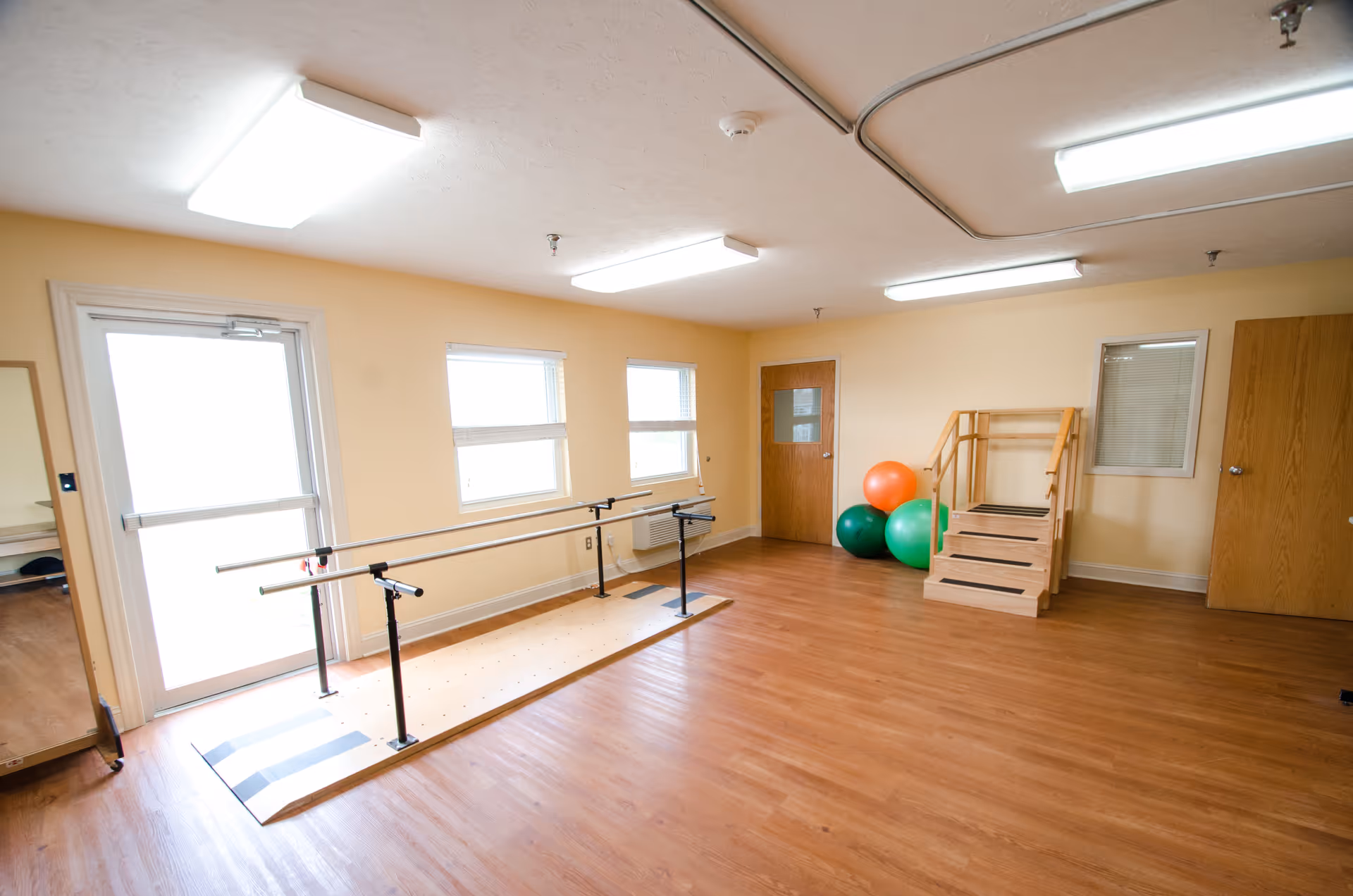 A spacious room with wooden flooring and beige walls, featuring parallel bars for physical therapy or rehabilitation exercises, a small wooden staircase with handrails, three large exercise balls in green and orange, three windows, two wooden doors, and a large mirror on the left side near a glass door.