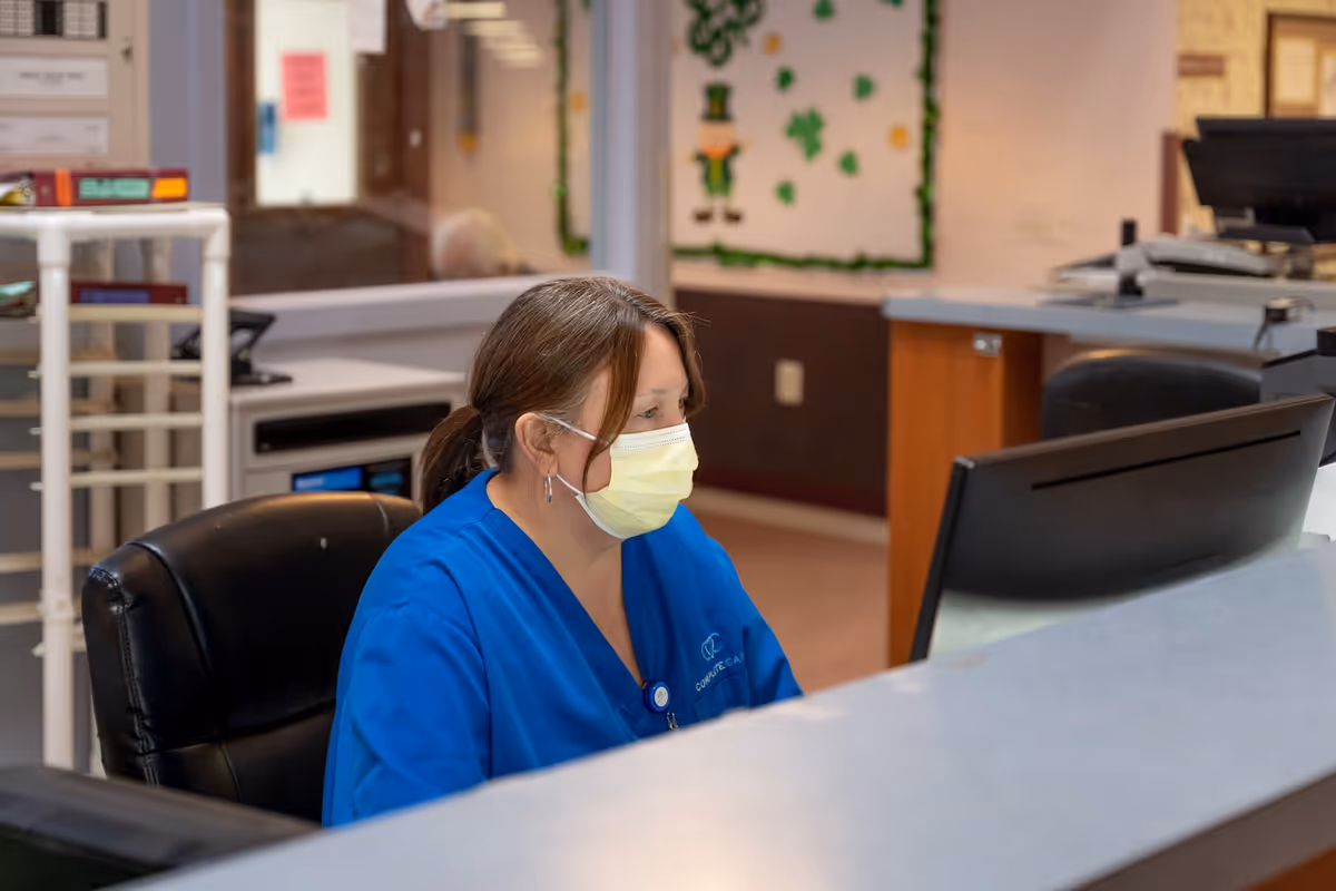 A healthcare worker wearing a blue uniform and a yellow face mask is seated at a reception desk, looking at a computer monitor in a care facility. The background shows a bulletin board decorated with shamrocks and a leprechaun, suggesting a St. Patrick's Day theme.