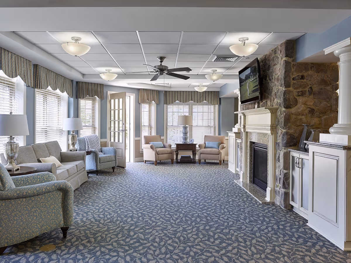 Sunlit communal lounge with sofas and armchairs, a stone fireplace with mounted TV, and large windows with blinds.
