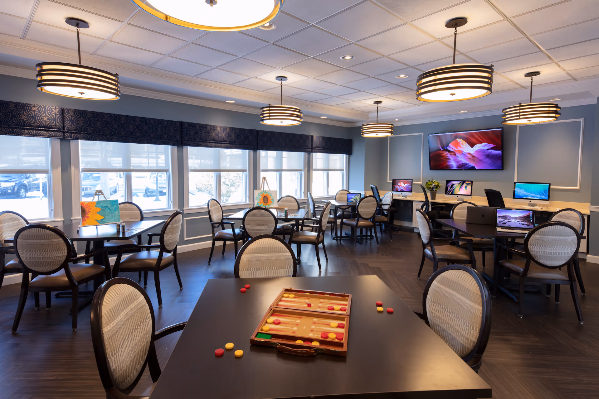 Bright communal activity room with tables and chairs, pendant lights, large windows, TVs and computers at the back, and games and paintings on the tables.
