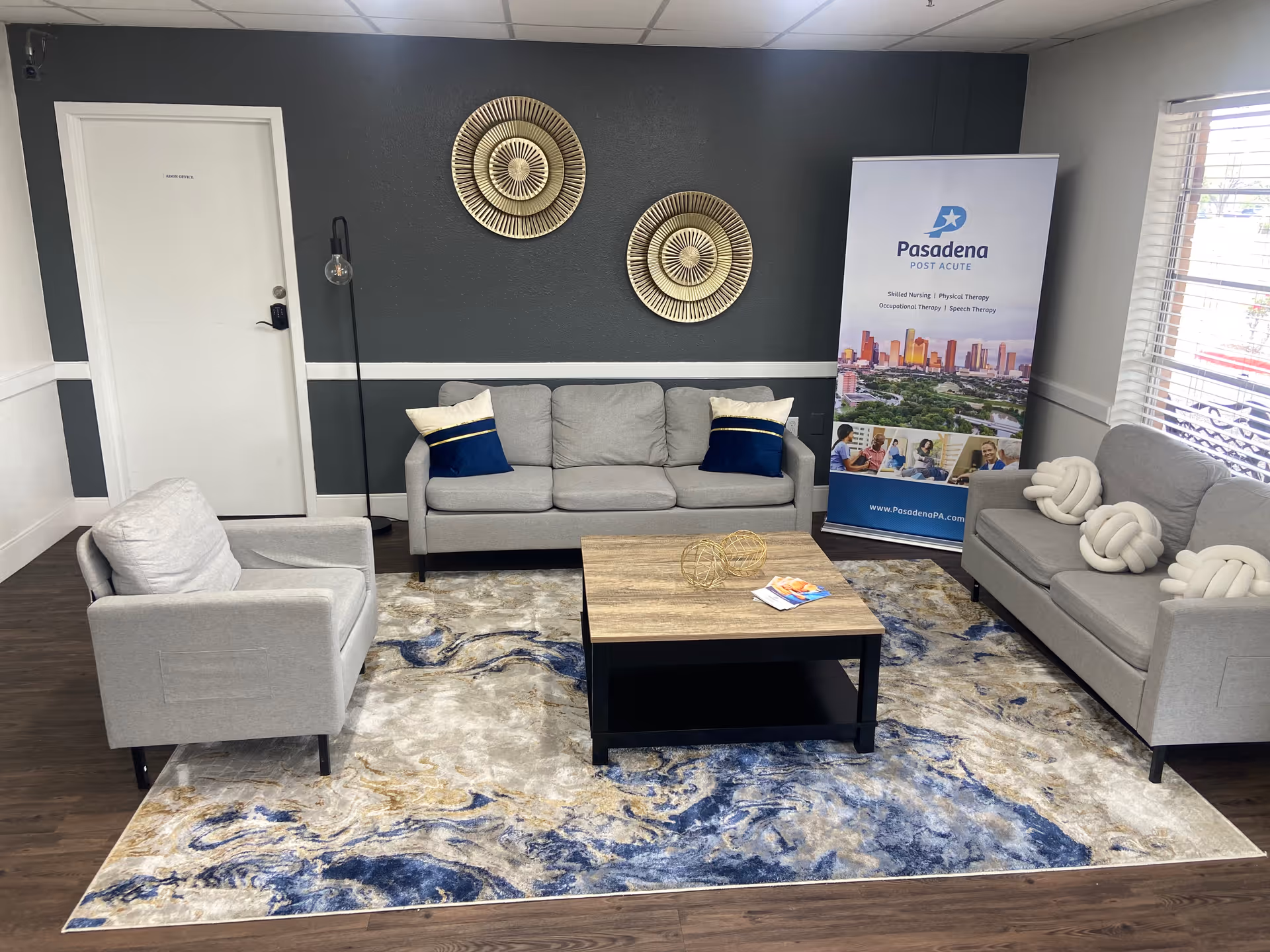 Reception lounge with gray sofas and armchairs around a wooden coffee table, decorative wall art, and a Pasadena Post Acute banner.