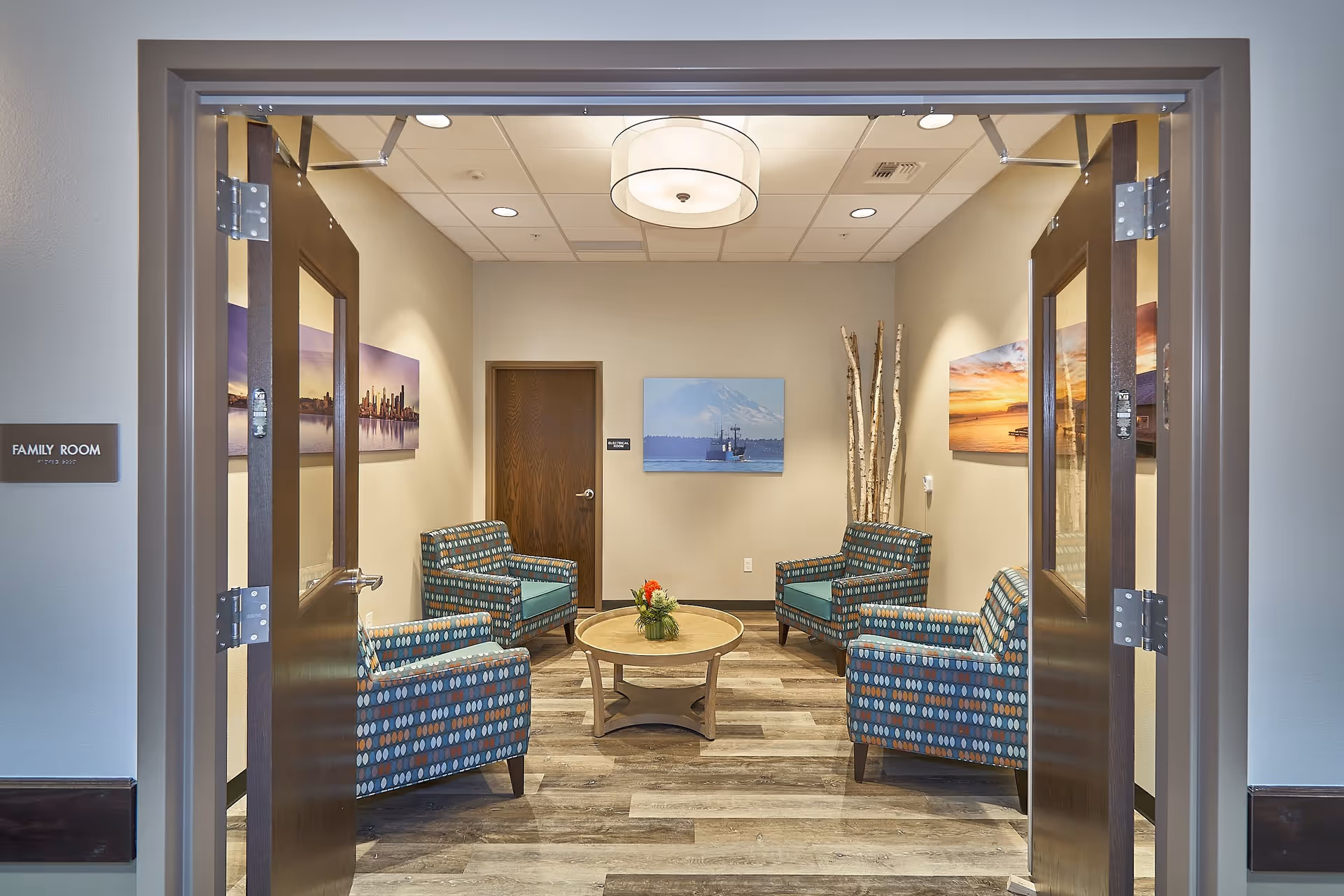 Interior view of a family room in a senior living facility with four patterned armchairs arranged around a round wooden coffee table with a small flower arrangement. The room has light-colored walls, wood flooring, a ceiling light fixture, and three landscape paintings on the walls. There is a closed wooden door at the back and a bundle of decorative birch branches in the corner.