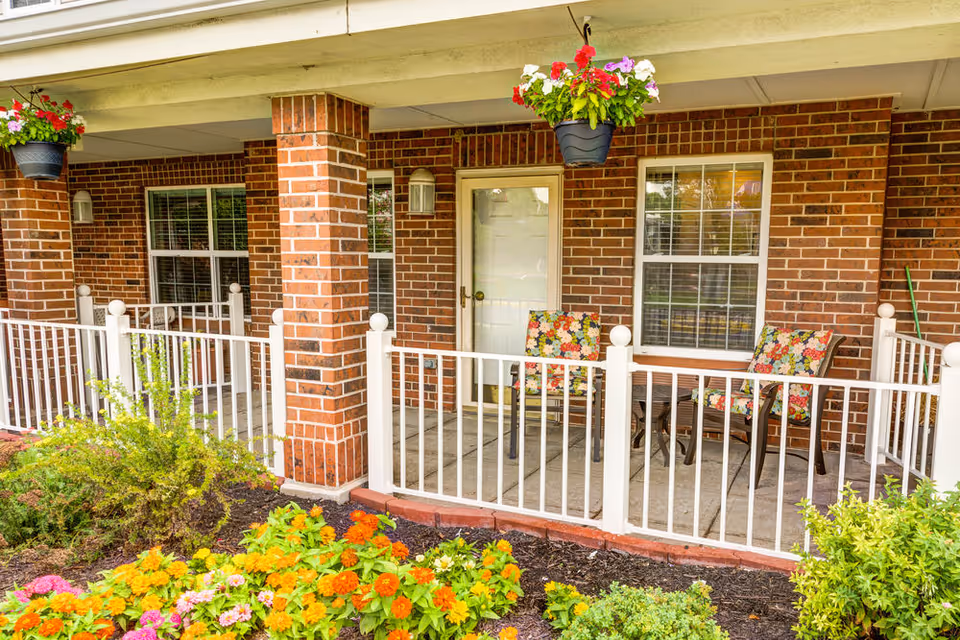 A covered outdoor patio area with two floral-patterned cushioned chairs and a small table between them. The patio is enclosed by a white railing and supported by brick columns. Hanging flower pots with colorful flowers are suspended from the ceiling. In front of the patio, there is a garden bed with vibrant orange, yellow, and pink flowers and green shrubs.