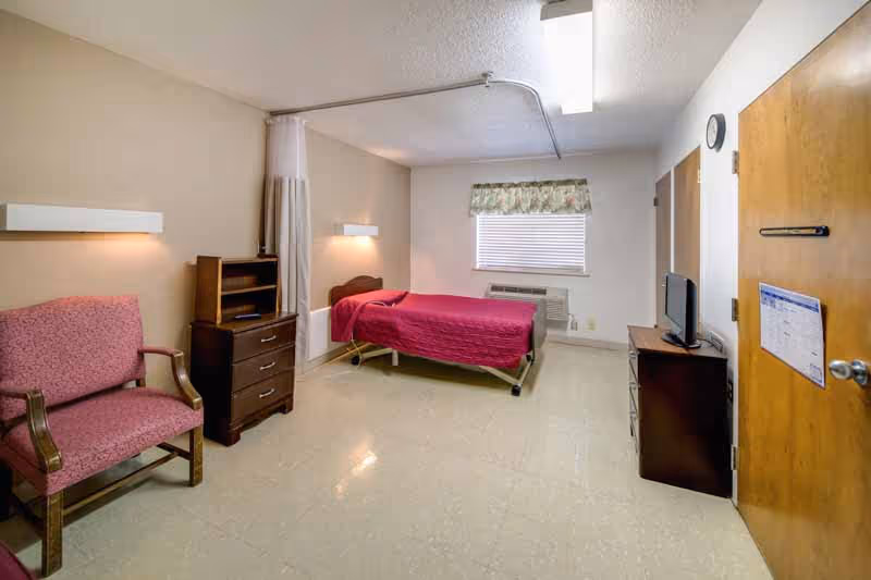 A simple, clean bedroom in a healthcare facility featuring a single bed with a red bedspread, a pink upholstered chair, a wooden dresser, a small TV on a stand, a window with blinds and a floral valance, and a door with a schedule or notice attached.