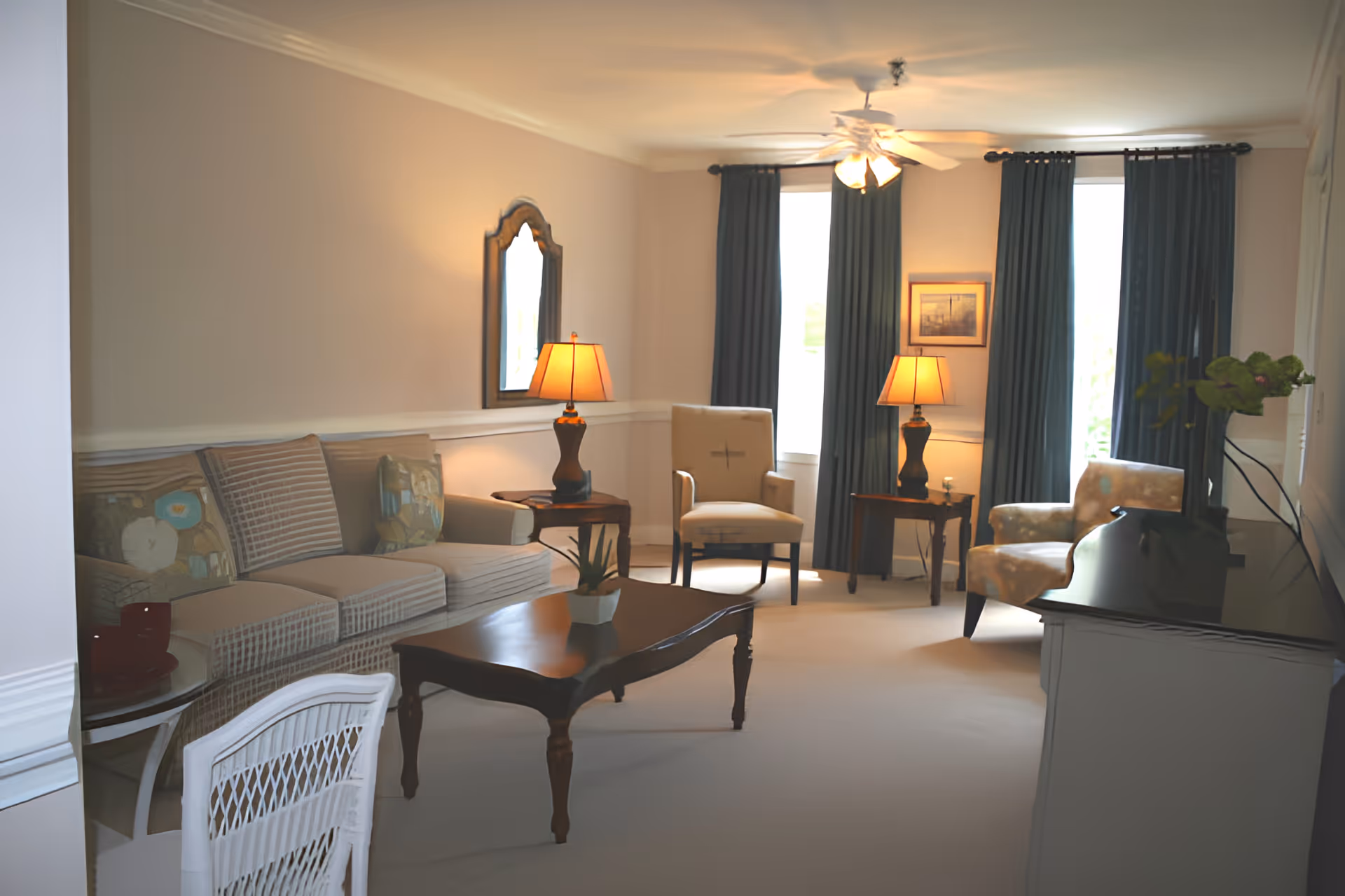 A cozy living room with a beige sofa adorned with patterned cushions, two armchairs, a wooden coffee table with a small plant, two side tables each with a lamp, a wall mirror, dark curtains covering windows, and a ceiling fan with lights.