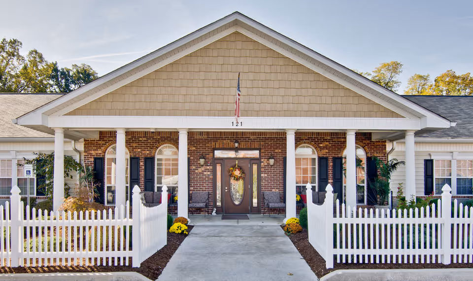 Front exterior view of a senior living facility named Chestnut Glen Senior Living with a white picket fence, brick facade, four white columns supporting a covered entrance, and a door decorated with a wreath. There are chairs on the porch and some plants and flowers around the entrance.