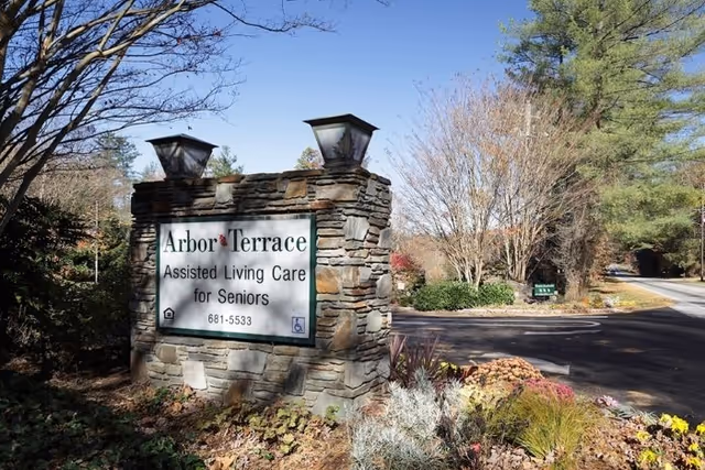Stone sign for Arbor Terrace Assisted Living Care for Seniors with two lanterns on top, surrounded by plants and trees along a paved road under a clear blue sky.