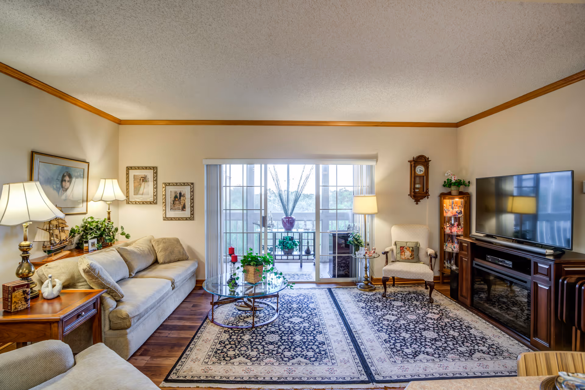 A cozy living room with a beige sofa, armchair, and glass coffee table on a patterned rug. The room has wooden flooring, a large flat-screen TV on a wooden stand, a grandfather clock, and framed pictures on the walls. Sliding glass doors lead to a balcony with plants outside.