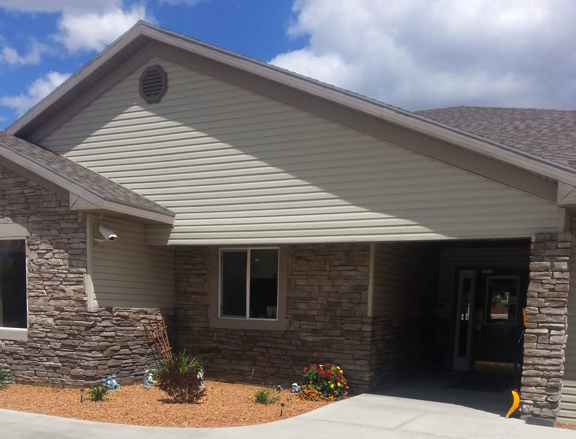 Front entrance of a single-story building with stone veneer and siding, a covered drive-through entrance, and small landscaped beds under a blue sky.