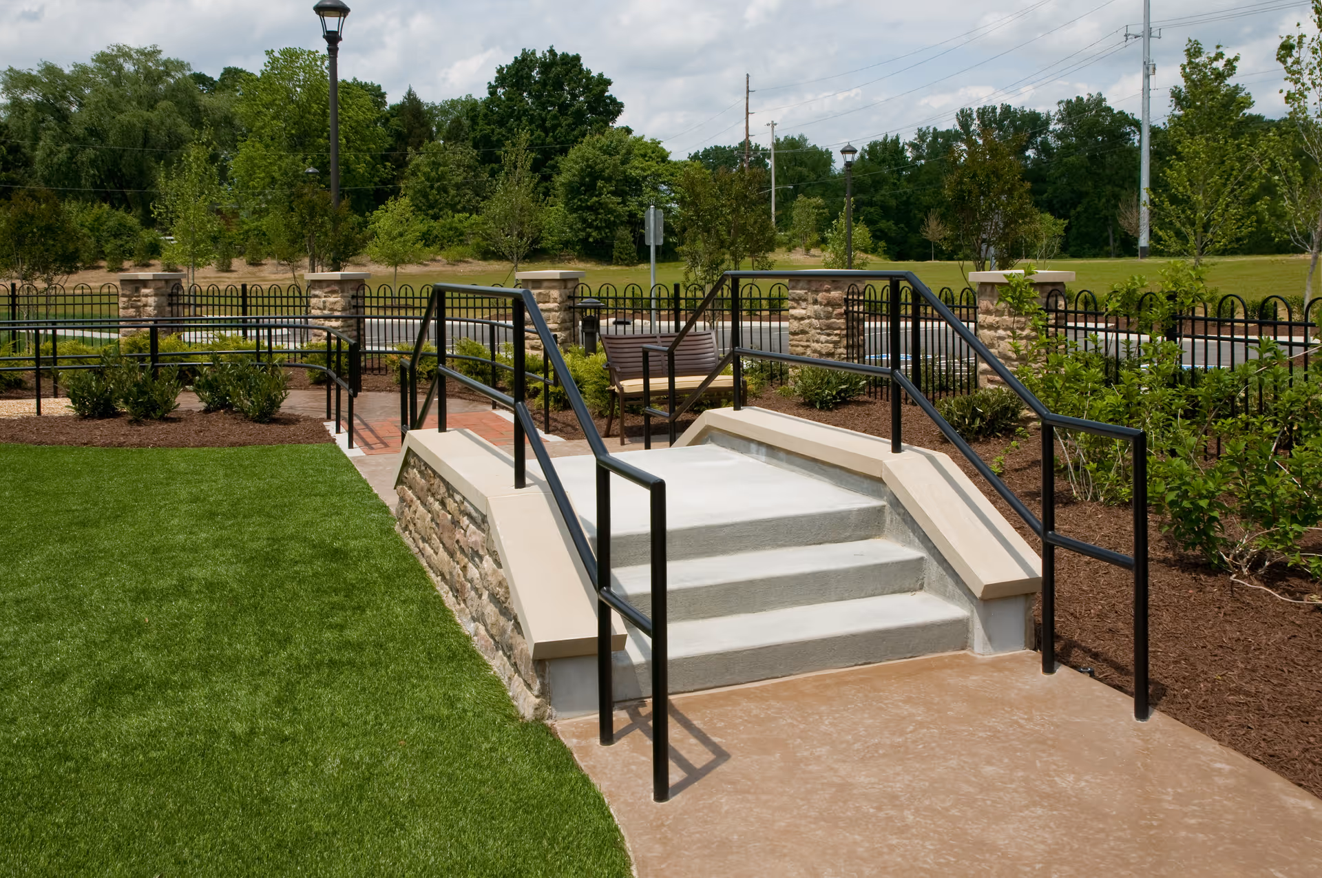 Landscaped outdoor courtyard with concrete steps and black metal handrails, a bench, lawn, and ornamental fencing.