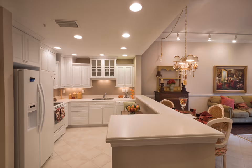 Bright open kitchen with white cabinets, a refrigerator, a long countertop/island, and an adjacent dining and living area with a chandelier and sofa.